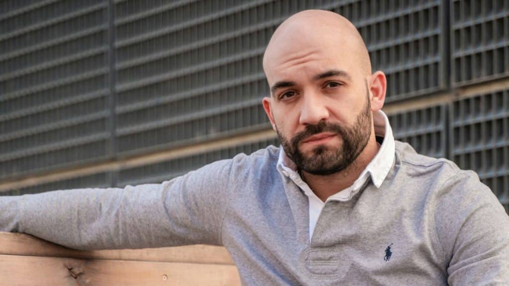 A bald man with a beard, wearing a gray polo shirt, sits looking directly at the camera.