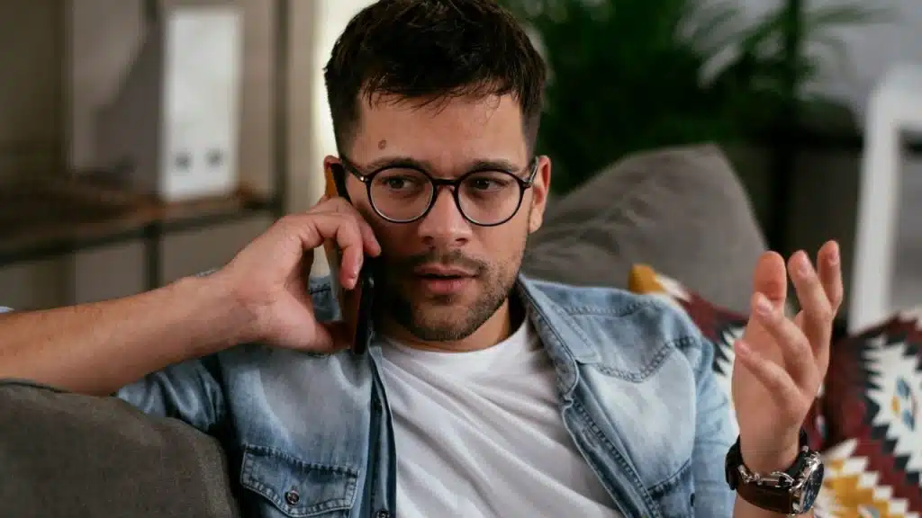 A man in glasses and a denim shirt sits on a couch talking on a cell phone.