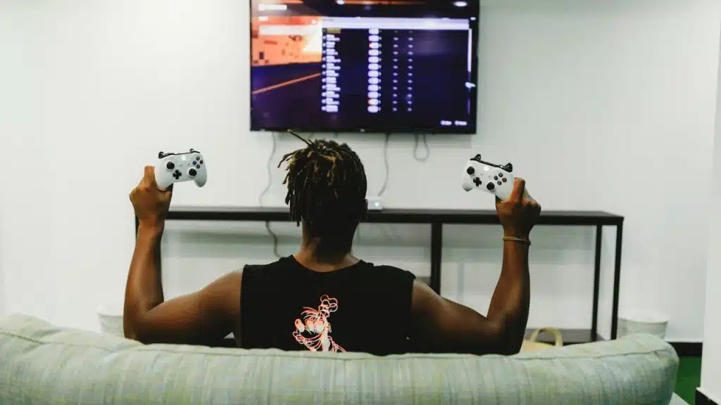 Man with dreadlocks holding two white controllers facing a TV screen.
