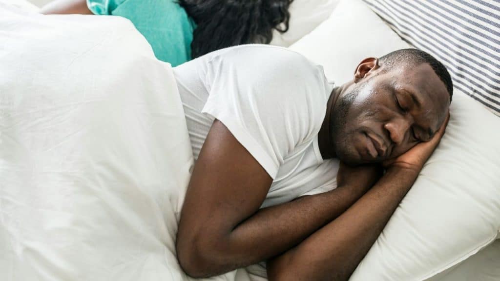Man in a white t-shirt sleeping on his side, woman partially seen in background.
