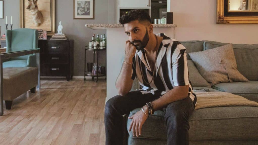 A bearded man in a striped shirt sits thoughtfully on a sofa in a living room.