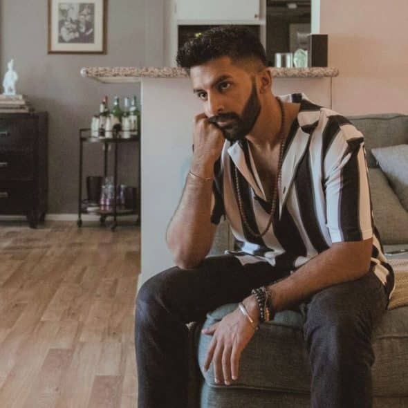 A bearded man in a striped shirt sits thoughtfully on a sofa in a living room.