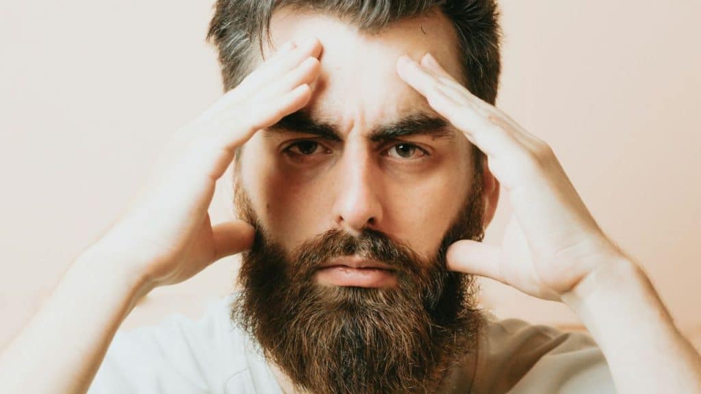 A man with a full beard and short hair holds his hands to his temples, looking stressed.
