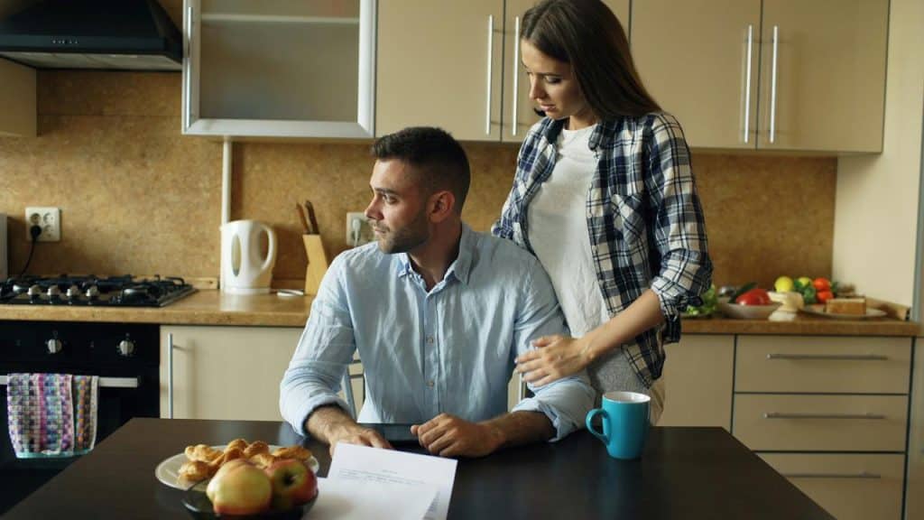 A woman standing beside a seated man in a kitchen, with food and papers on the table.