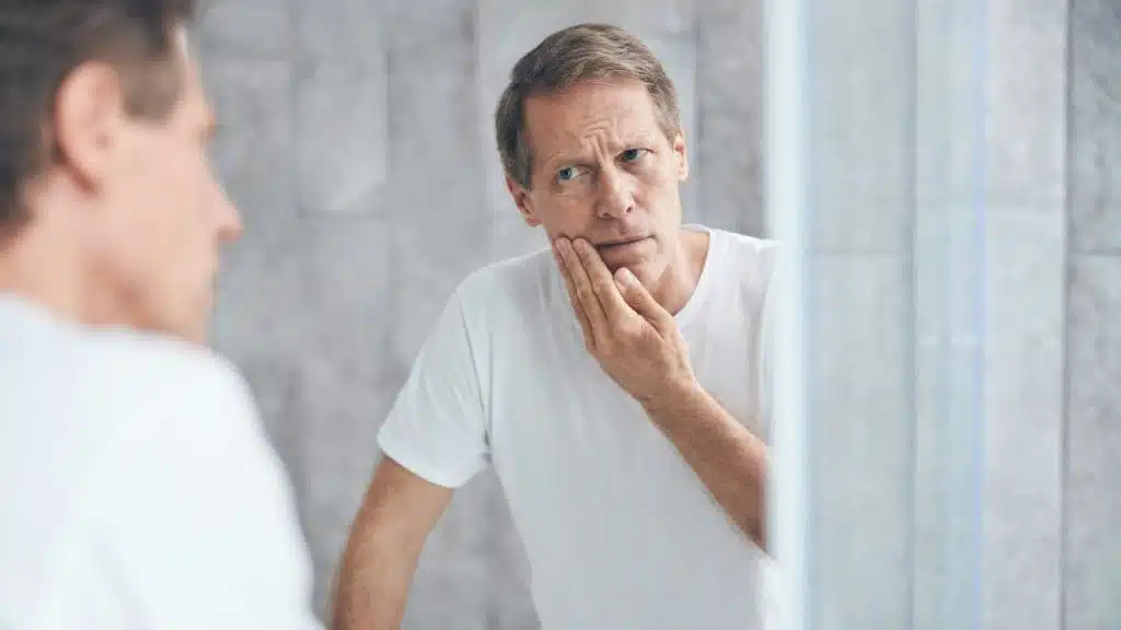 A middle-aged man looks intently at his reflection in a bathroom mirror.