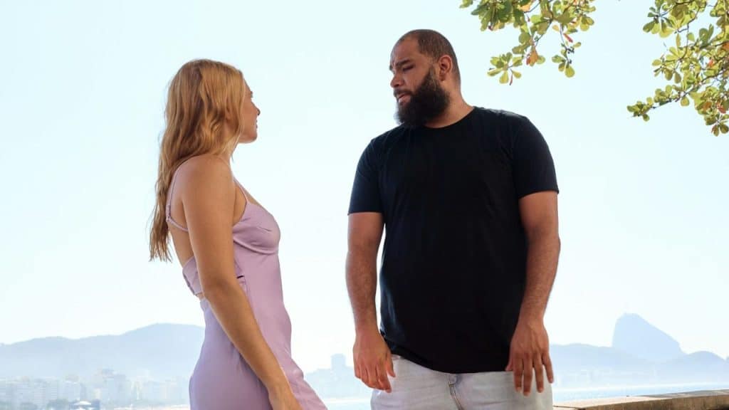 A blonde woman in a lavender dress talking to a bearded man outdoors near the coast.