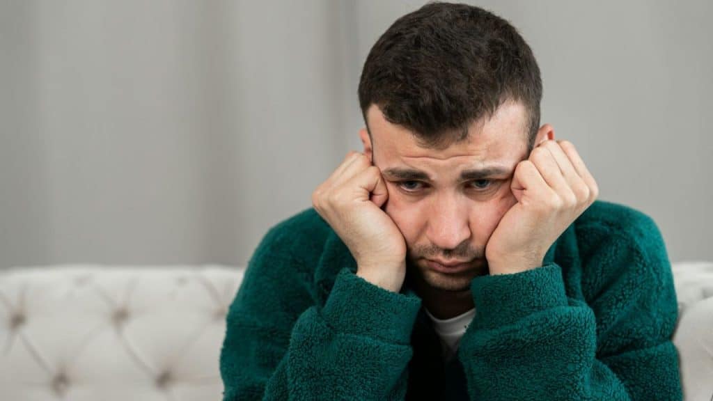 Man with worried expression, hands on cheeks, looking down in a green sweater.