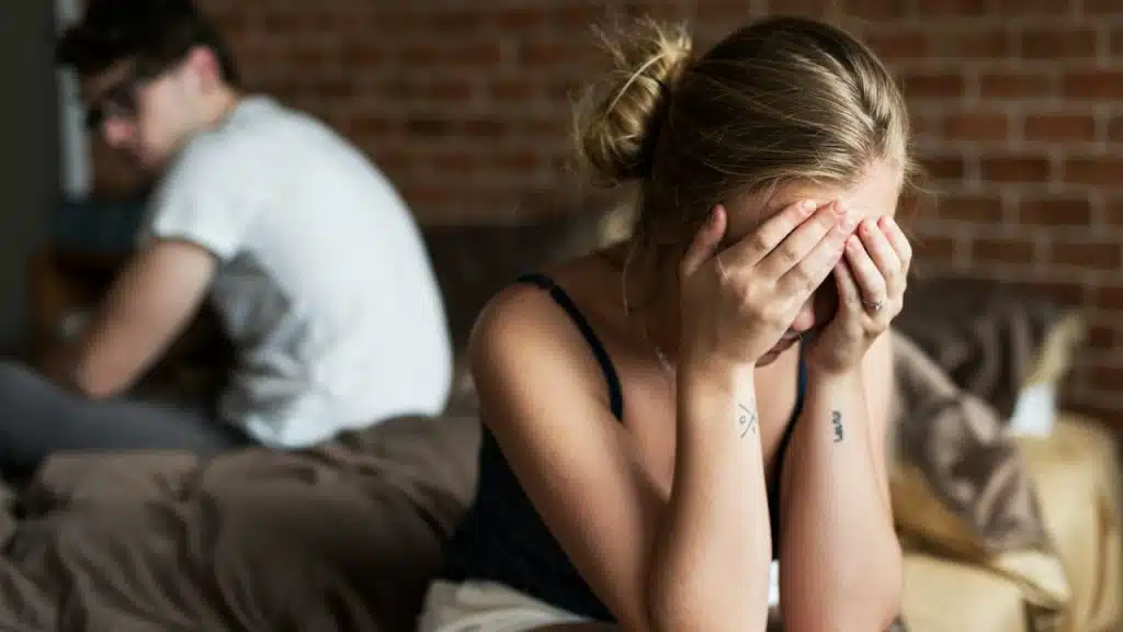 A woman sits distraught with her hands covering her face while a man sits blurred in the background.