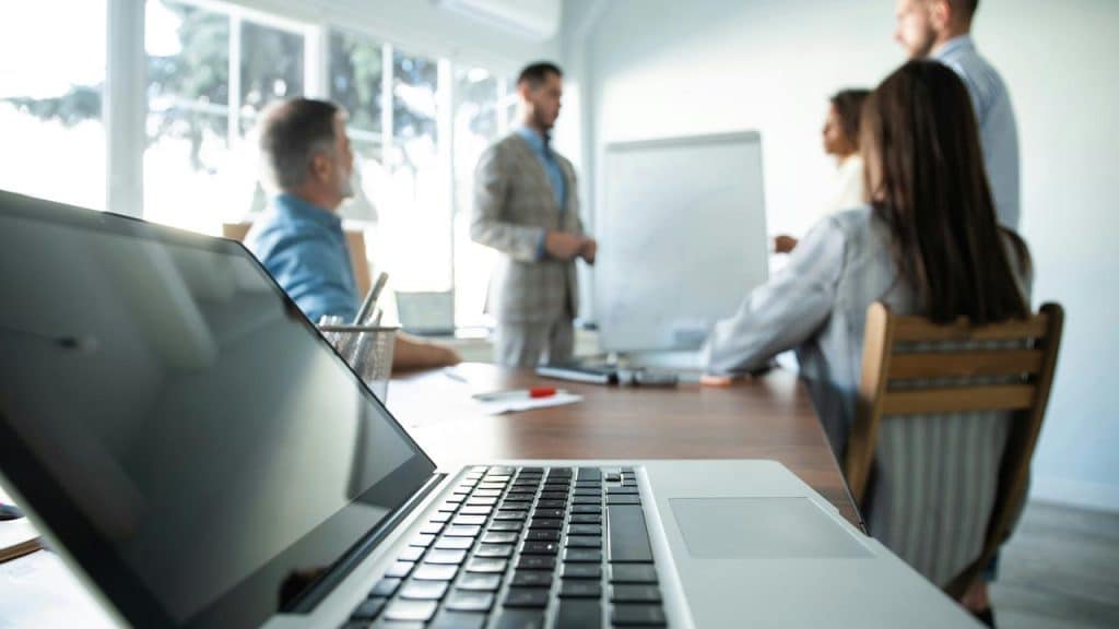 A laptop in the foreground as a team gathers around a whiteboard for a meeting.