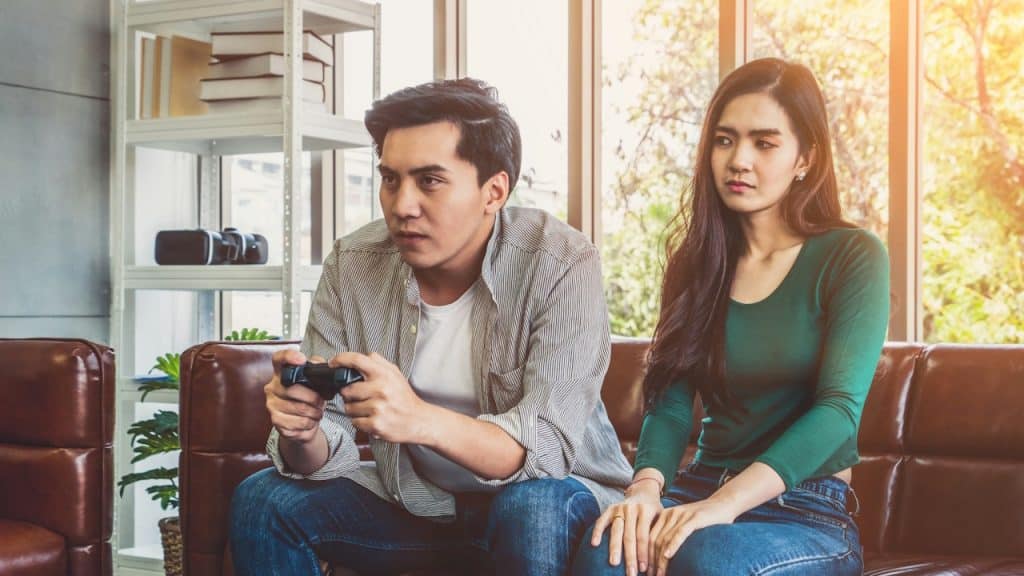 A man is intensely focused on playing a video game with a controller, while a woman sitting next to him on the couch looks annoyed.