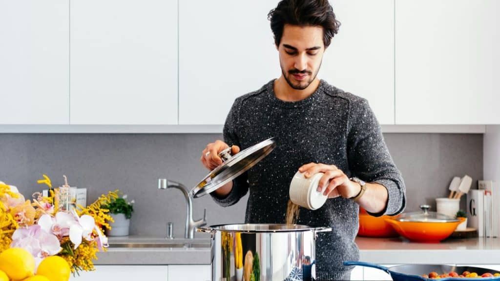 Man in a grey sweater adding seasoning to a pot in a modern kitchen.