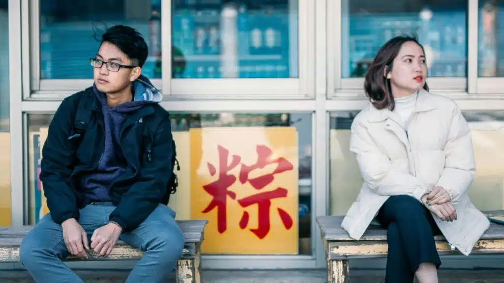 A man and a woman sit outdoors on benches, looking away from each other; a yellow sign with a large red character is between them.