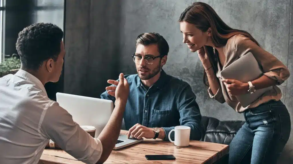 Two men and one woman discussing work around a laptop at a wooden table.
