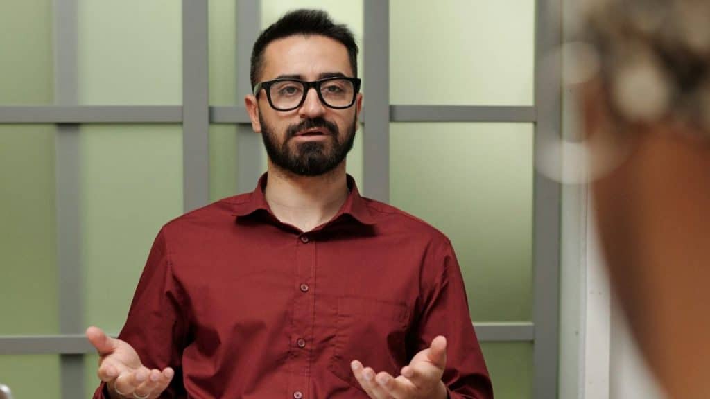 Man with a beard and glasses in a maroon shirt gesturing to a person nearby.