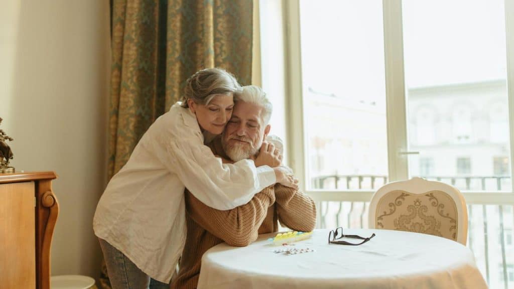 A senior woman hugging a seated senior man at a table near a window.