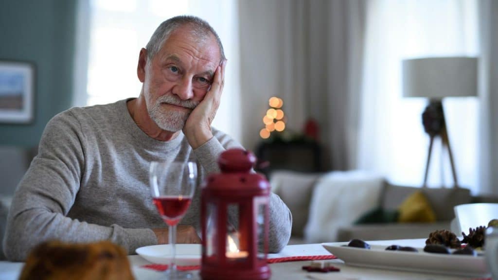 An older man with a gray beard sits alone at a table, resting his head on his hand.