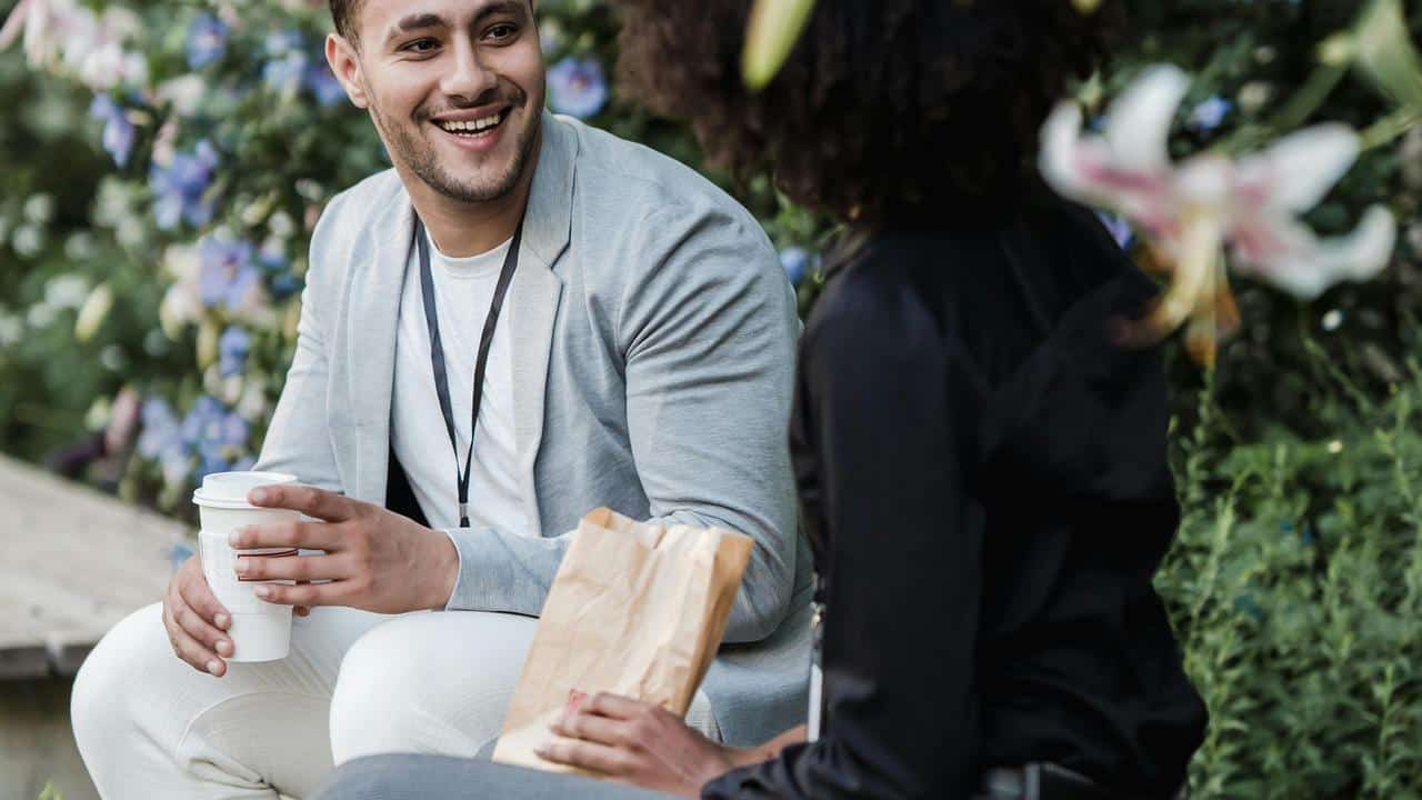 A man smiling and talking with a woman outdoors while holding a coffee cup.