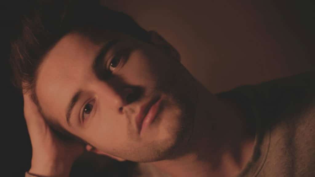 A man in a dark sweatshirt sits in a dimly lit room looking directly at the camera.