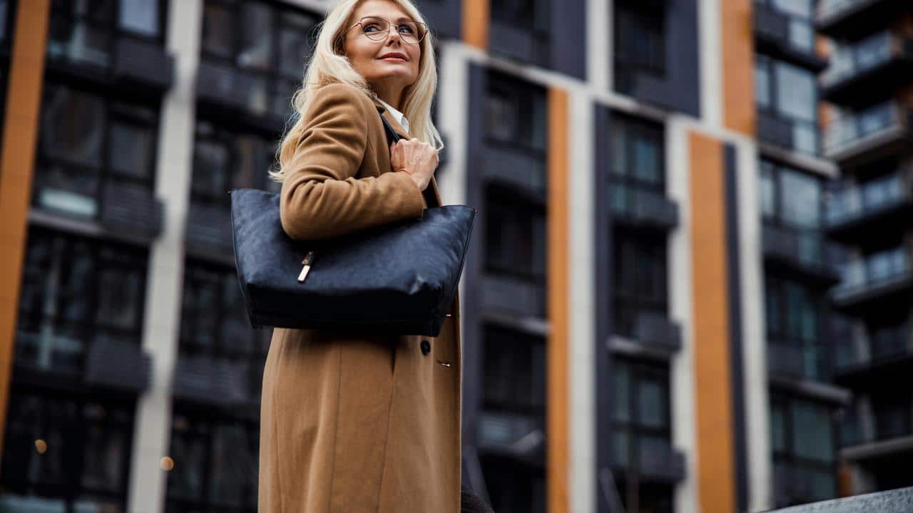 A woman in a brown coat holding a black bag while standing outside modern buildings.