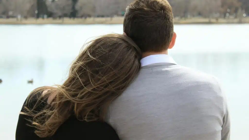 A couple sitting by the water with the woman resting her head on the man’s shoulder.