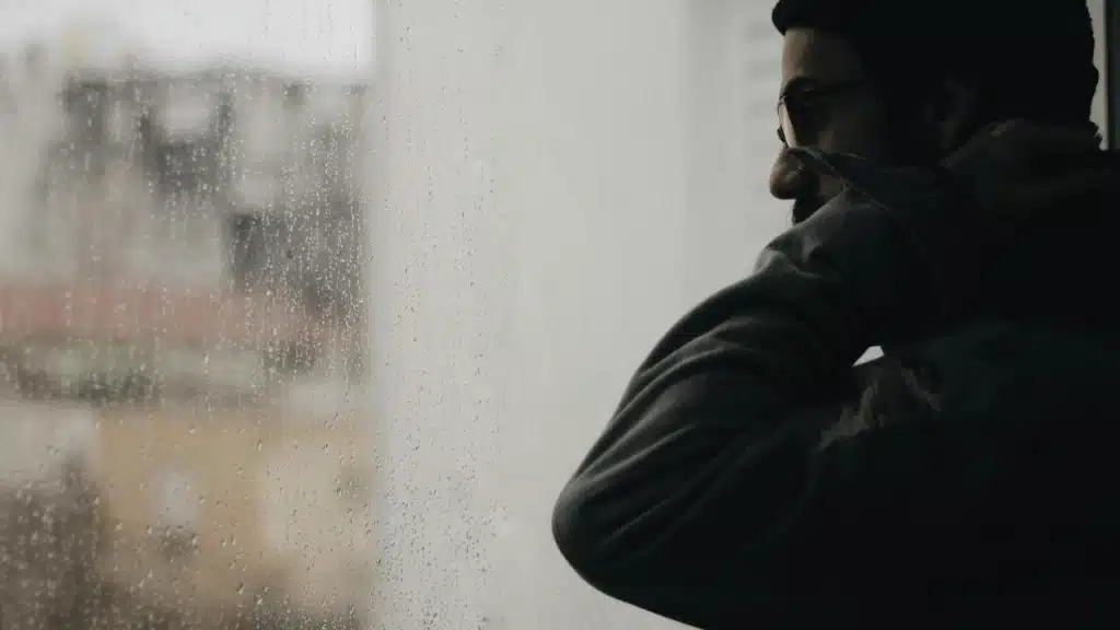 A person in profile standing by a rain-speckled window, looking outside thoughtfully.