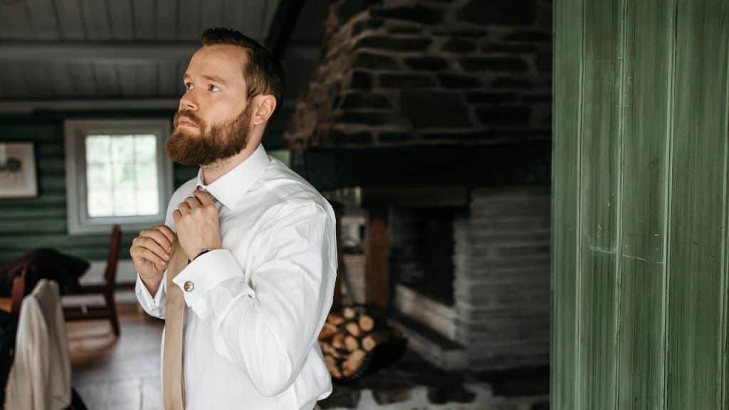 A bearded person adjusting a tie inside a rustic room.