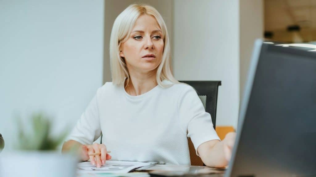 A blonde woman in a white shirt sits at a desk and looks at a computer screen.