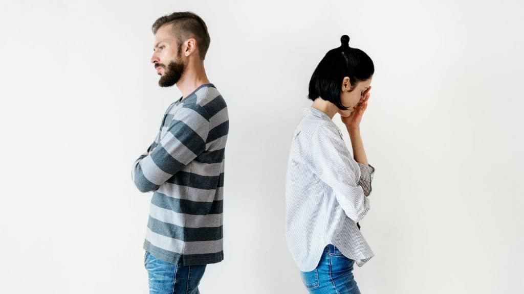 A bearded man stands with crossed arms while a woman bows her head against a wall.