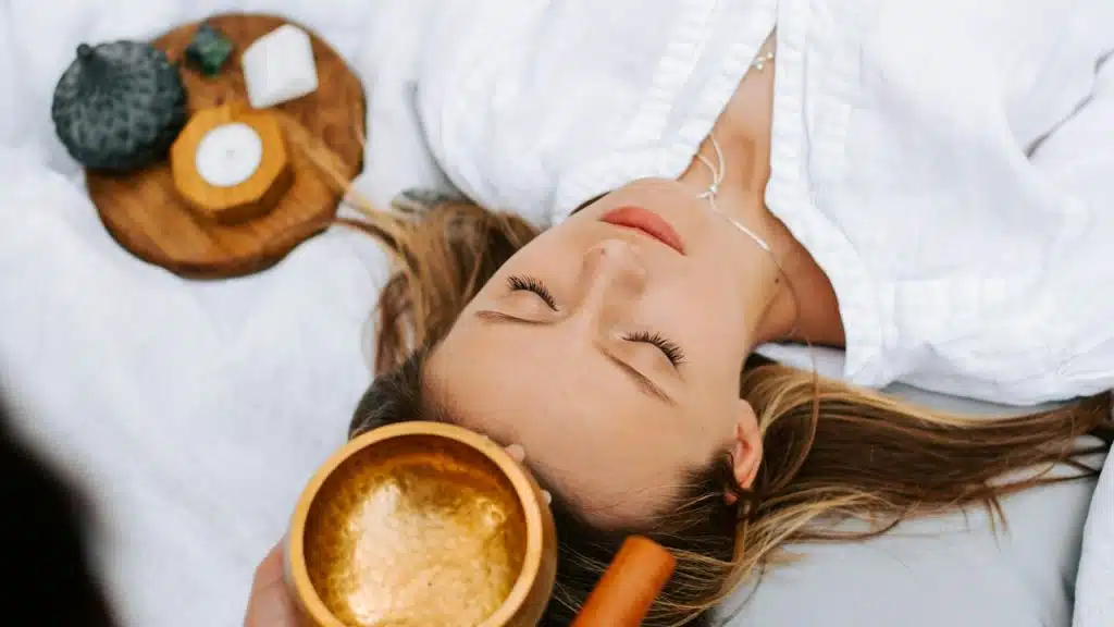 A woman lying on a bed with closed eyes while someone stands over her holding a bowl of containing a liquid.