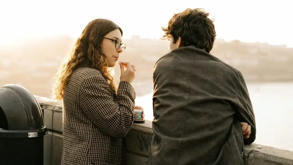 A woman eating ice cream and looking at a man while they stand by a barrier overlooking a lake.