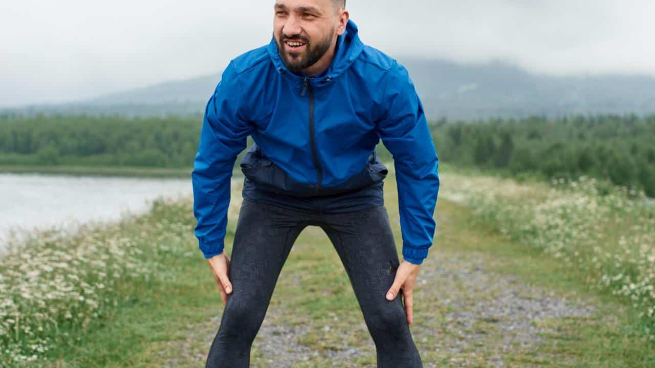 A man in a blue jacket resting after a run on a nature trail.