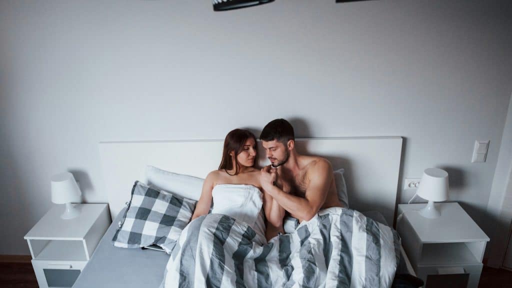 A couple wrapped in blankets sitting up in bed, looking affectionately at each other.