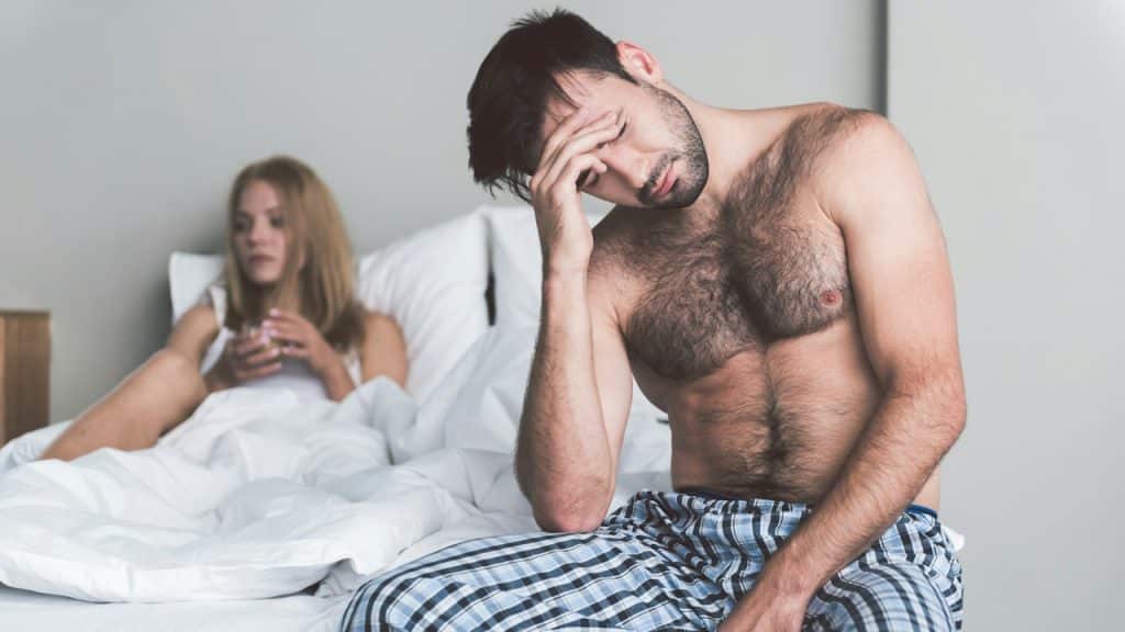 A shirtless man sitting on a bed, looking distressed with his hand on his head, and a woman in the background.