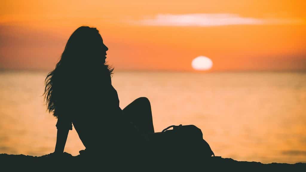 The silhouette of a woman sitting on the ground with a bag beside her, looking out at the sun setting over the ocean.