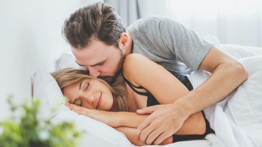 Man hugs and kisses a sleeping or resting woman in bed under white covers.