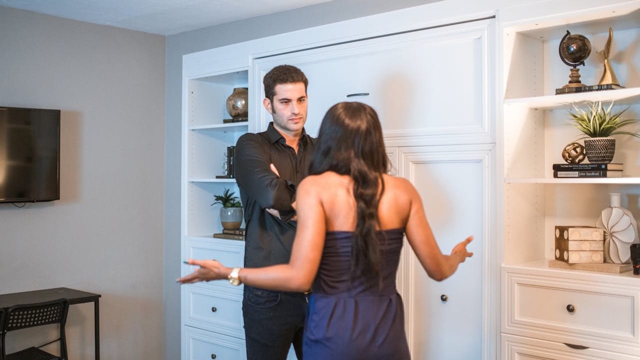 A couple arguing in a room, with the man standing with his arms crossed and the woman gesturing towards him.