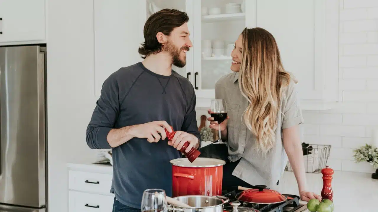Couple smiling at each other in a bright white kitchen, cooking and holding wine.