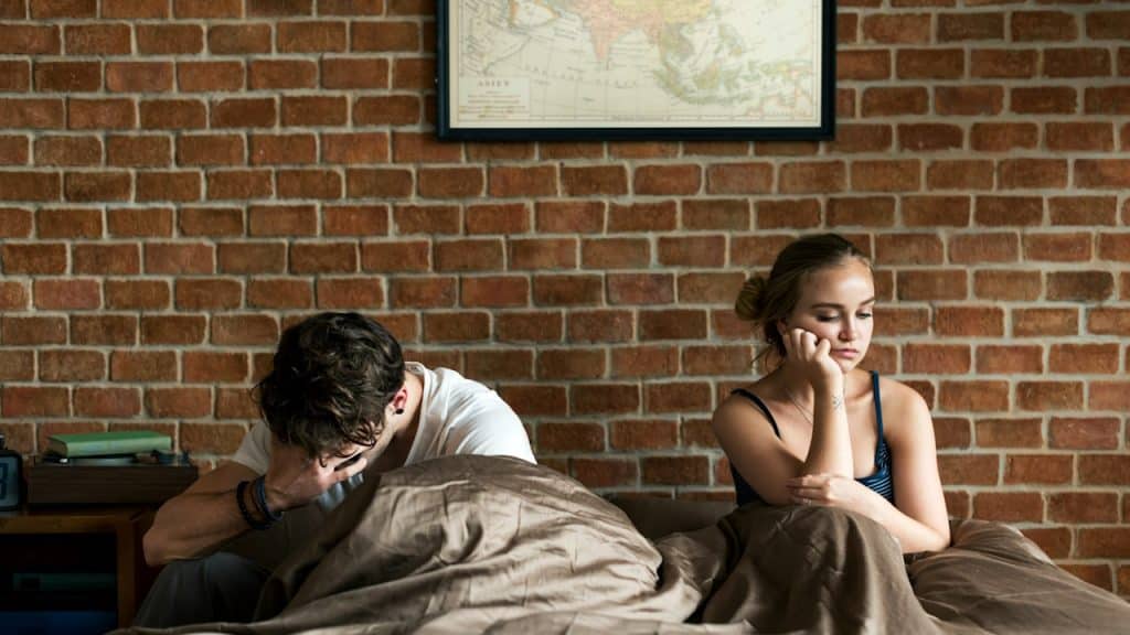 A young man is touching his head and closing his eyes with desperation; a sad woman is sitting in bed along him.