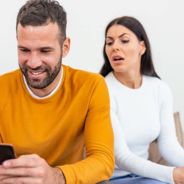 A smiling man looking at his phone while a suspicious woman peers over his shoulder.