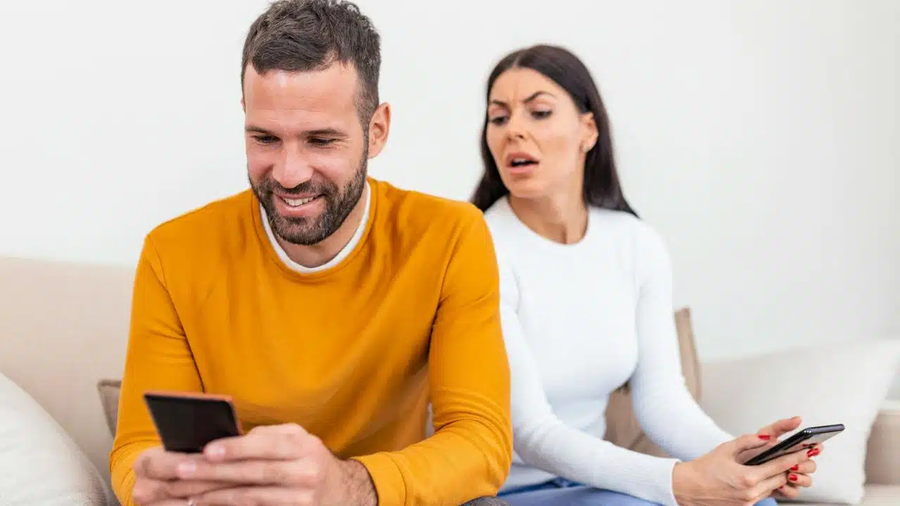 A smiling man looking at his phone while a suspicious woman peers over his shoulder.