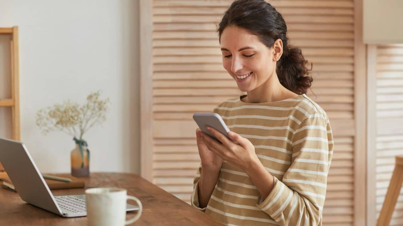 A smiling woman in a striped shirt looks at her phone while sitting at a wooden table with a laptop and coffee mug.