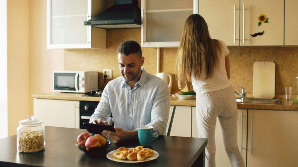 A man uses a tablet at a table while a woman works in the background kitchen.