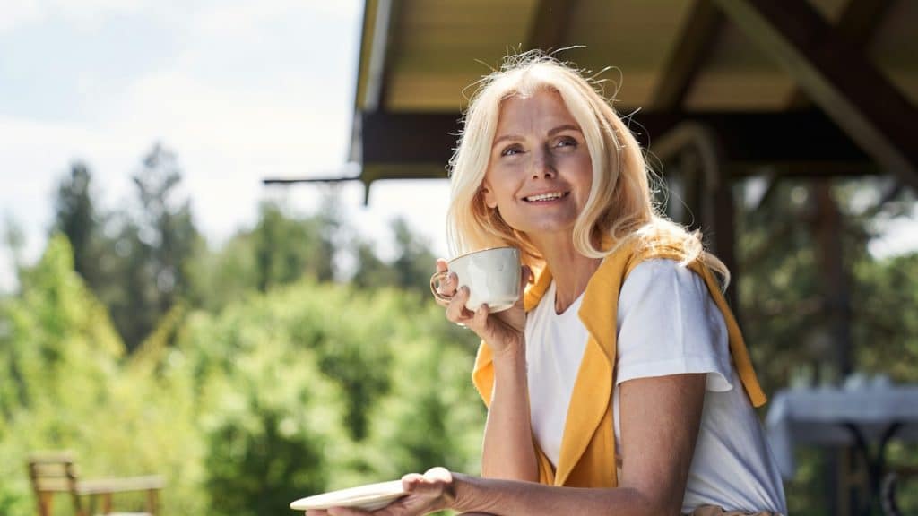 A smiling, attractive female sitting on the terrace of a countryside house and having a cup of coffee.