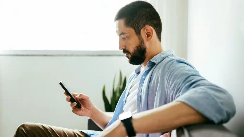 A bearded man looking at his phone’s screen while sitting on a couch.