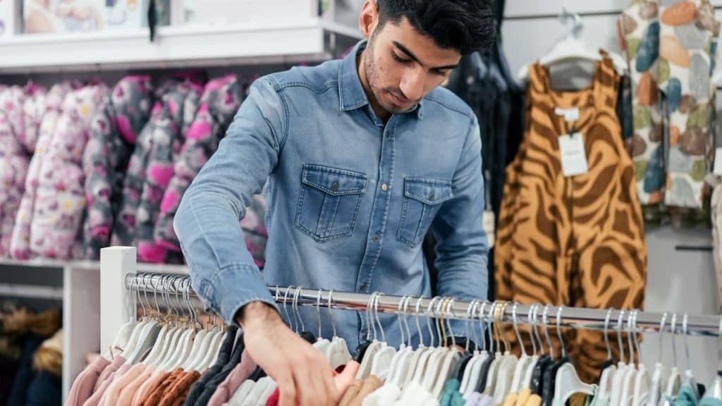 A man in a denim shirt examining clothes on a rack in a retail store.