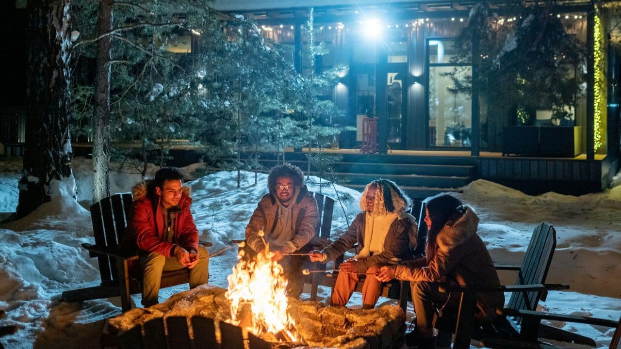 A group of four people sit around a large bonfire in winter snow at night, roasting marshmallows.