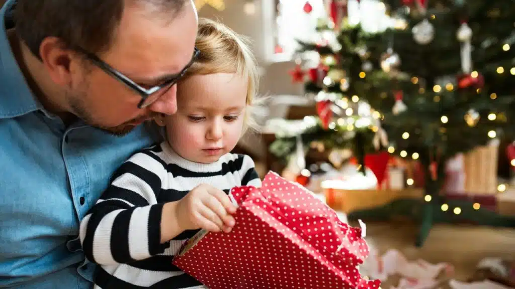 Father wearing glasses helping child open a red, polka-dot gift near a Christmas tree.