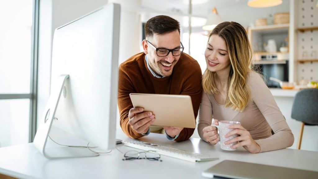 Smiling man and woman looking at a tablet together while sitting at a white desk.