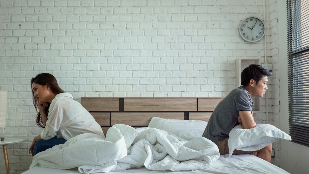 A man and woman sitting on a bed with their backs to each other, looking away after an argument.