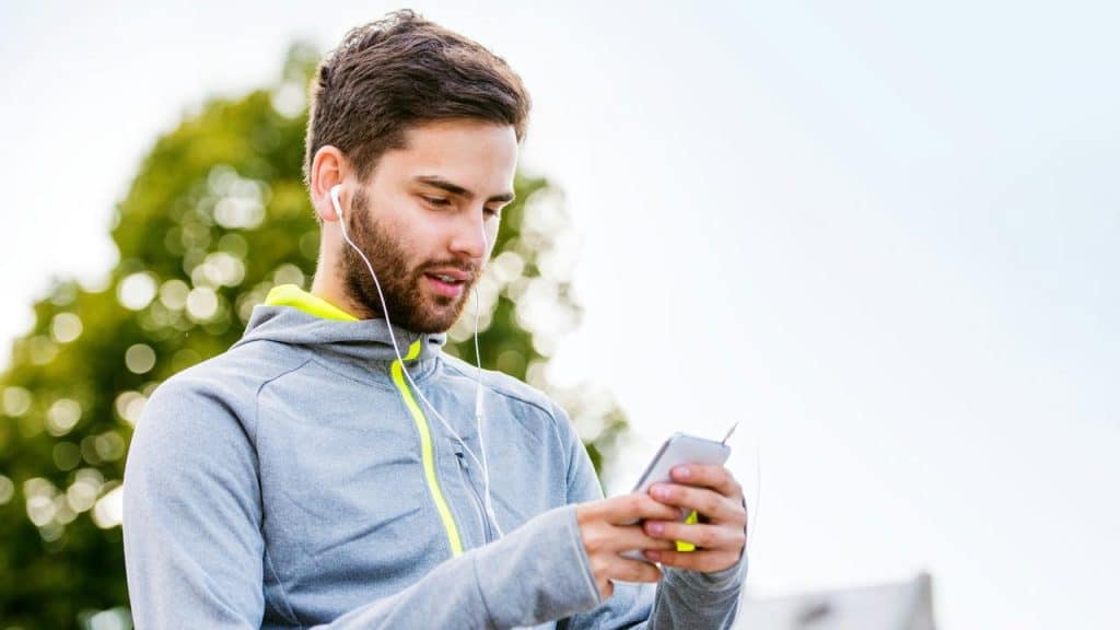 A person wearing earphones and a gray hoodie using a smartphone outdoors.
